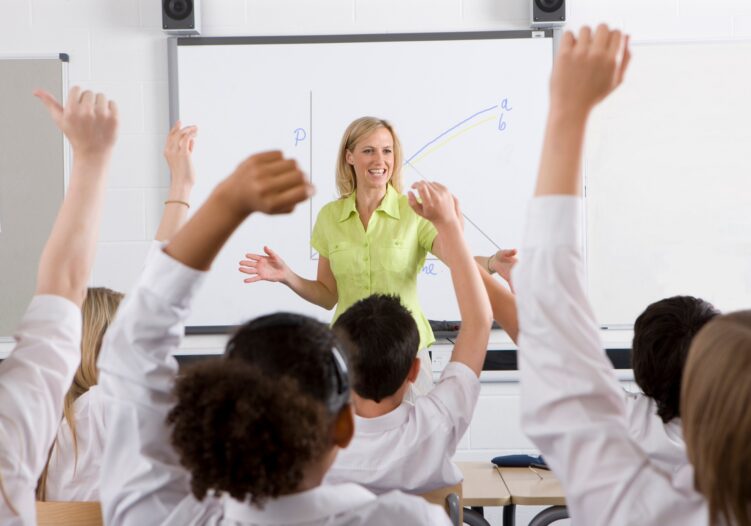 Rear view of class, students with their hands raised and Teacher stood at the front